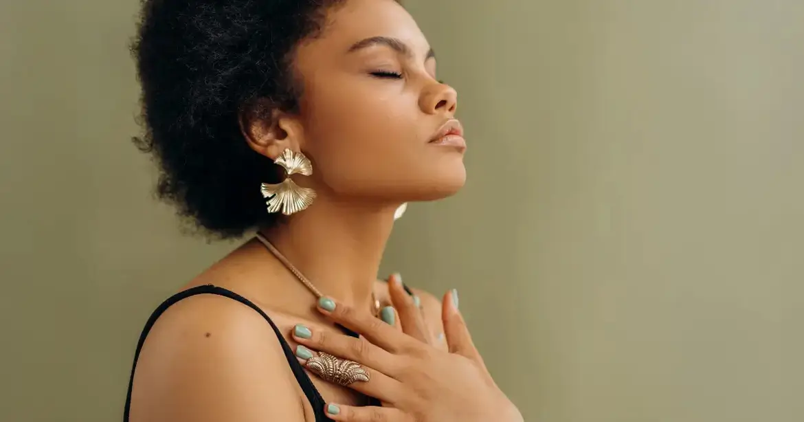 A person with short curly hair and large earrings is standing against a neutral background. They have their eyes closed and hands placed gently on their chest, wearing a ring and a necklace.