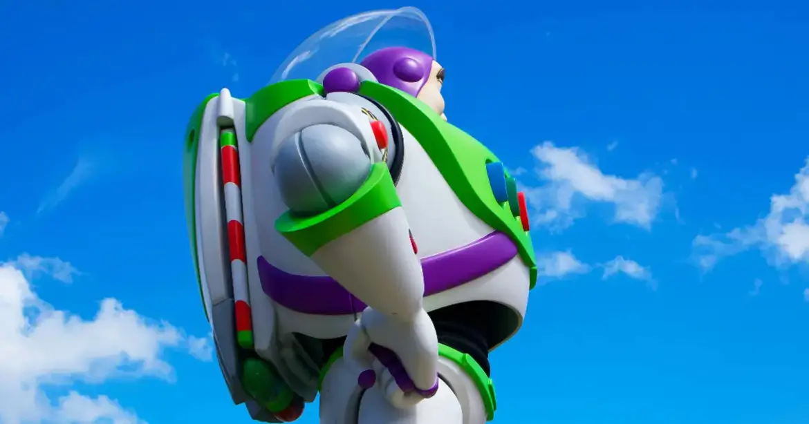 A large statue of a space-themed toy character with a green and white suit, purple accents, and a clear helmet stands against a bright blue sky with a few clouds. The character is looking...