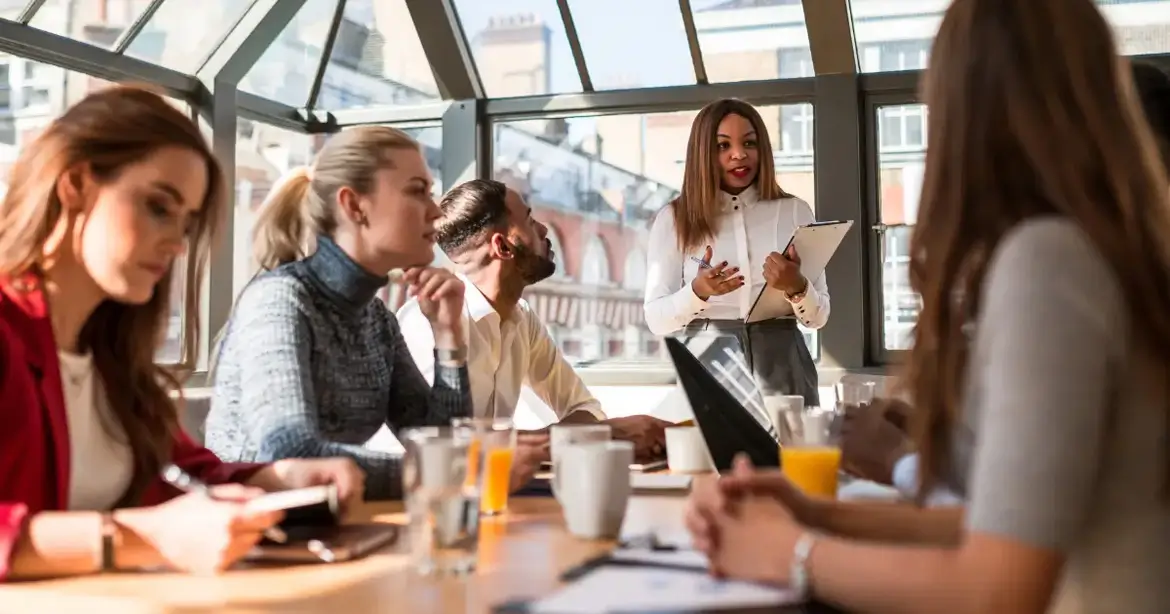 A group of people sitting around a table in a bright, modern conference room. A woman standing and holding a clipboard is speaking to the group. The table is filled with notebooks, glasses of...