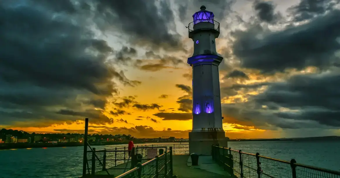 A lighthouse on a pier at sunset, with a dramatic sky filled with dark clouds. The lighthouse is illuminated with blue lights, and the horizon glows with orange and yellow hues. The sea...