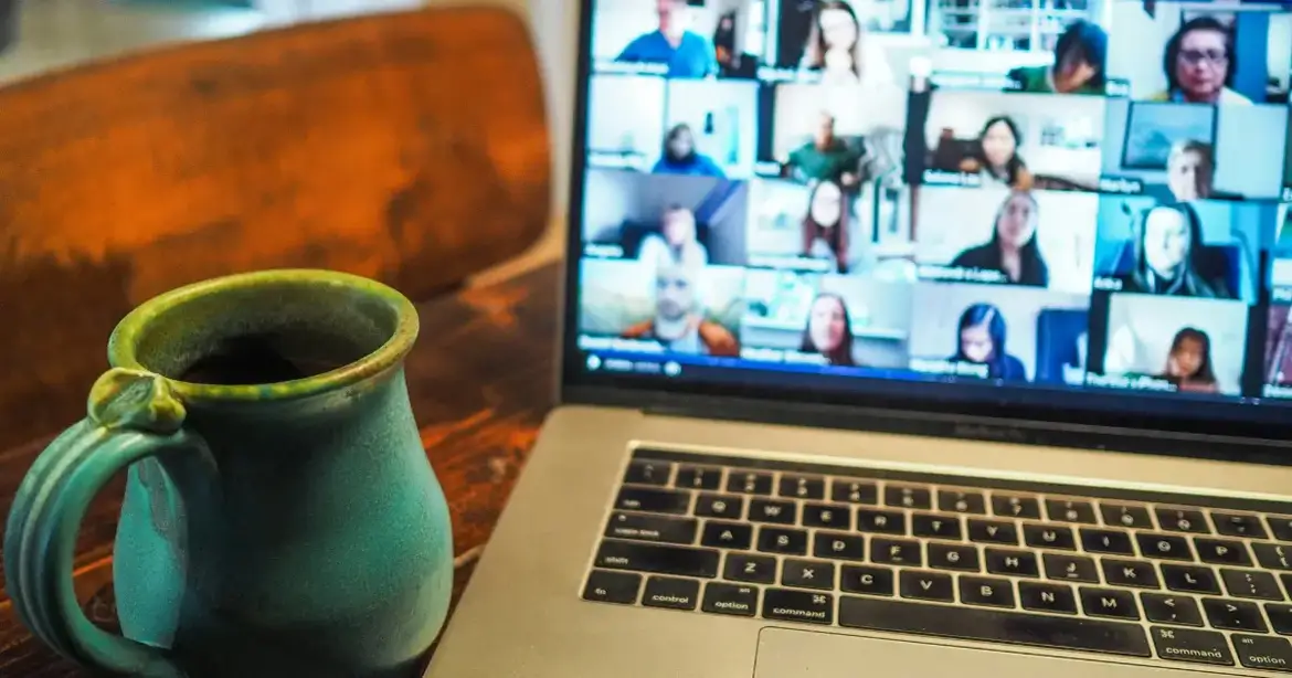 A laptop displaying a video conference with multiple participants in a grid layout. A green ceramic mug is placed on the wooden table next to the laptop.