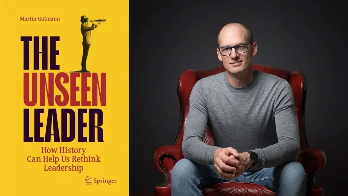 A man wearing glasses and a gray sweater sits in a red armchair, holding a book titled "The Unseen Leader: How History Can Help Us Rethink Leadership" by Martin Gutmann. The book cover is...