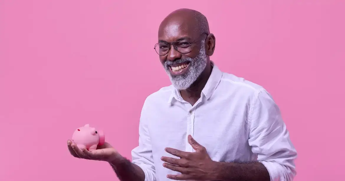 A smiling man with a beard and glasses, wearing a white shirt, holds a small pink piggy bank against a pink background.