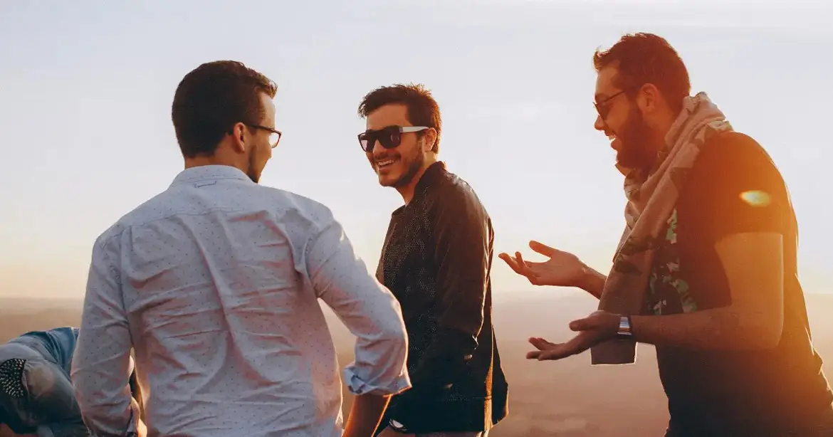 Three men are standing outdoors, engaged in healthy conversation and smiling. They are casually dressed, and the sun is setting in the background, casting a warm glow.
