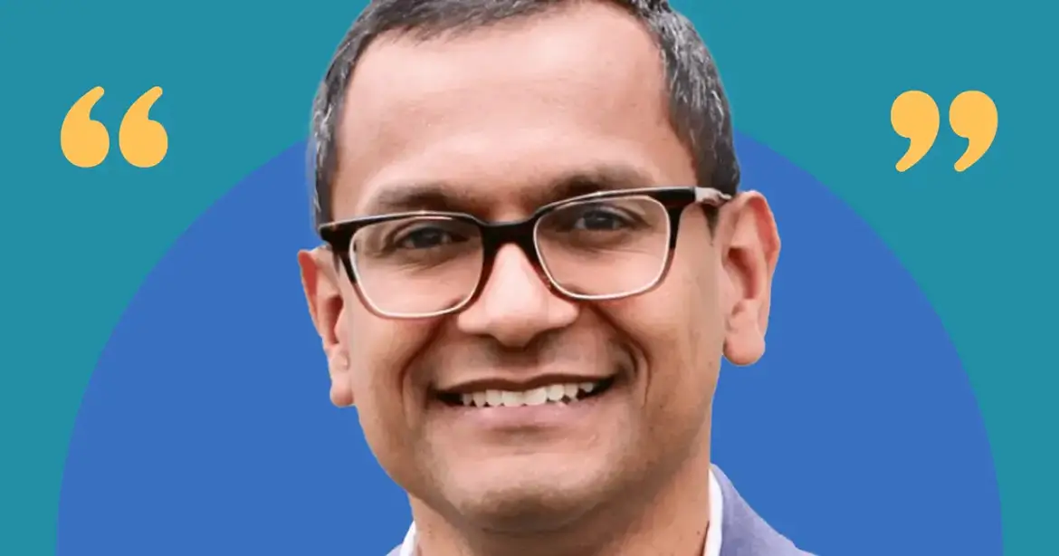 A person wearing glasses and a suit, smiling against a blue background with yellow quotation marks on either side.